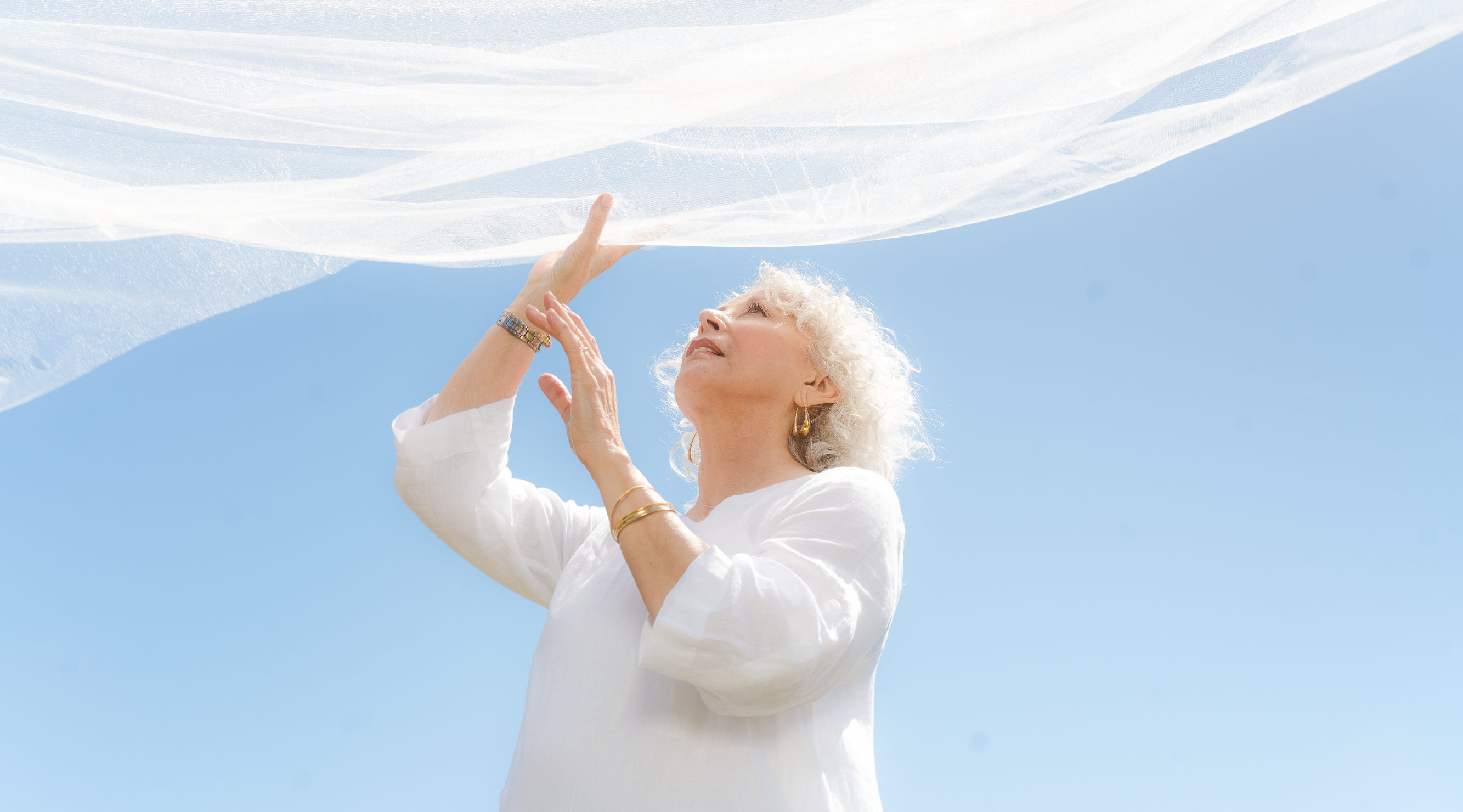Nevaeh Day Spa. Woman with grey curly hair wearing all white reaching up to wispy white fabric flowing in the wind with blue skies behind her.