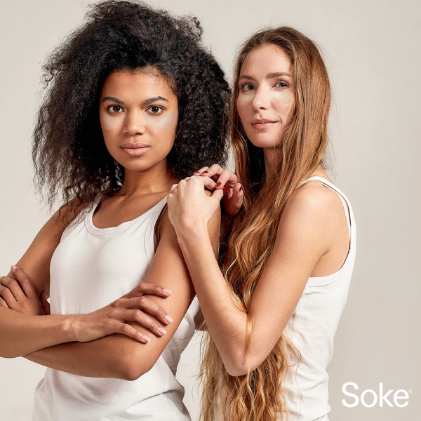 Two women standing with white tank tops on using the SOKE eye masks.
