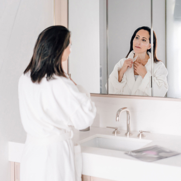 SOKE Neck treatment: image shows a woman wearing a white robe looking the in mirror about to apply the mask.